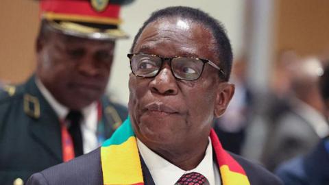 A head and shoulders image of Emmerson Mnangagwa wearing a Zimbabwe scarf around his next and a pair of glasses. He is looking to the left of the picture.