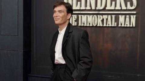 Cillian Murphy stood with his hands in his pockets wearing a long black blazer with a white shirt and striped black trousers