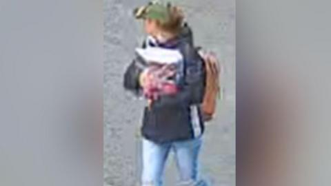 A grainy CCTV image of a woman looking away from the camera, wearing a green cap, brown rucksack and brown jeans. She's carrying an unidentifiable object in her arms.