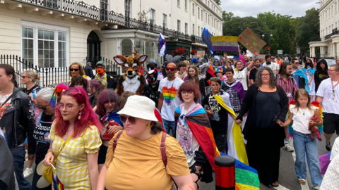 Crowds pass through the town centre. The march is about 8-10 people wide with many holding or waving Pride flags. There are men and women and one child is visible in the photo. Someone dressed as a fox is also walking in the parade.