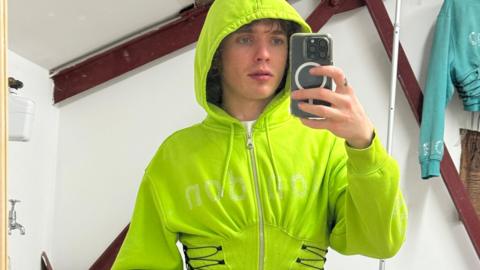 Timothy Gibbons wearing a lime green corset hoodie, he is holding up an iPhone