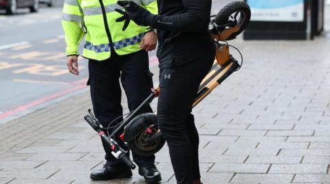 A police officer is talking to someone on a pavement as they hold a personal electric scooter