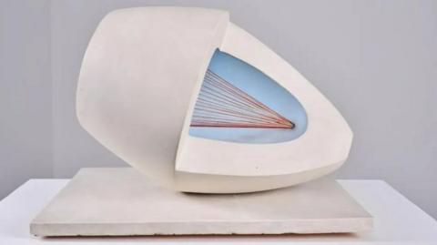 A white oval sculpture with pale blue and red colours in the middle. It has been placed on a white slab for display. 