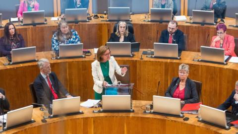 A group of Senedd members sat in the Senedd debating chamber, with the first minister Eluned Morgan stood at the front in the centre.