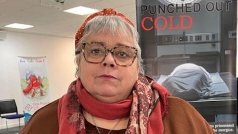 Maxine Thompson-Curl in front of a Punched Out Cold poster which has an image of a covered body in a morgue. She is looking pensive and wearing a headscarf, red scarf and brown jumper. 