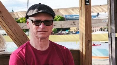 A man who wears a red t-shirt, sunglasses and a black flat cap sits in a wooden seating area. The background features wooden fencing and a waterfront with boats and buildings in the distance.