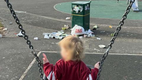 A child sat on a swing in a park. Infront of the child is an overflowing public bin and litter on the floor.