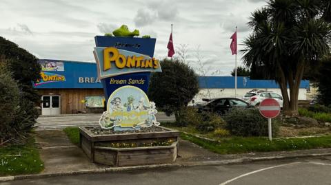 The exterior of Pontins Brean Sands, taken on a grey overcast day. It has a blue and yellow sign with a crocodile peering over the top, and there are two flag posts in the background. There is a small car park in front of the low single-story building. 