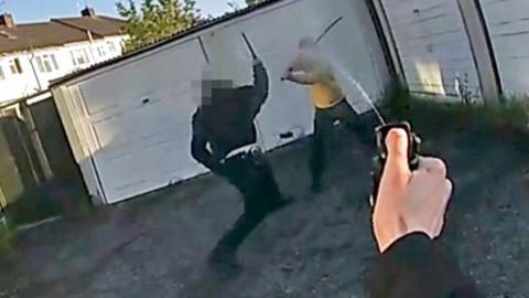 A still from police bodycam footage of a police officer holding up a baton as he confronts a man holding a long curved sword and wearing a yellow hooded top. The arm of the officer who is wearing the camera can be seen spraying pepper spray