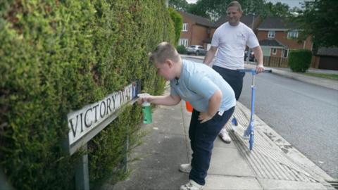 A young boy cleans a “Victory Way” street sign while his dad stands behind holding a scooter on a quiet suburban street.