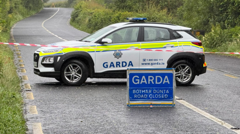 Shows a Garda car in white and yellow and a Garda sign saying road closed and translated into Irish.