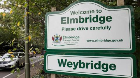 A sign for Elmbridge on the side of a road.