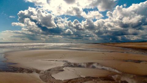 The sun shines through large white clouds in the blue sky over the beach exposed by receeded sea water