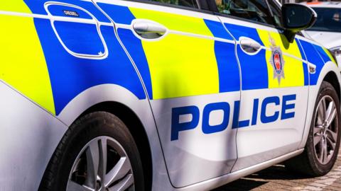 The outside of a police car which has yellow and blue square patterns on it and the word police as well as the Surrey Police crest