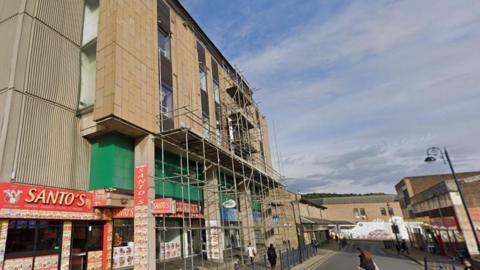 A street lined with buildings. On the left side there is scaffolding erected around a row of shops. There are people walking in the street and the sky is blue. 