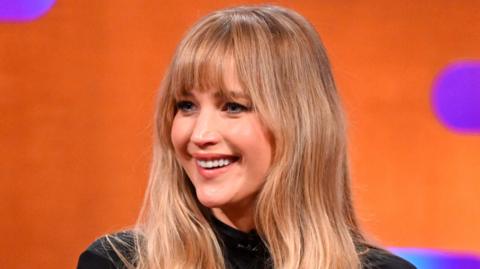 Jennifer Lawrence smiles in the Graham Norton Show studio. She has long brown hair and wears a black top