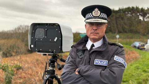 Robin Smith, Jersey Chief of Police, is positioned slightly to the right of the centre of the frame and is looking directly at the camera with a serious expression. He has short, greying hair and is wearing clear-rimmed glasses. He is dressed in a dark grey police uniform jacket with a high collar, epaulets, and "POLICE" patches on both shoulders. He also wears a traditional peaked police cap bearing the Royal Crest badge. His arms are folded across his chest. To the left of the man is a large, grey and black speed camera or surveillance unit mounted on a tripod. The camera unit is roughly rectangular, facing the same direction as the Robin's gaze, and is the dominant object in the background immediately behind him.