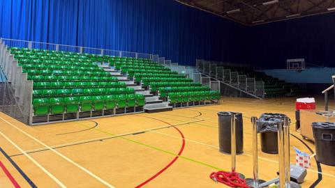 A sports hall with rows of green plastic seats and equipment on a wooden basketball court