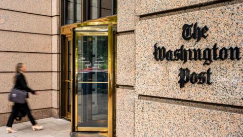 A woman dressed smartly in office attire walks towards the entrance of the Washington Post building. The words 'The Washington Post' are edged on the right-hand side of the entrance.