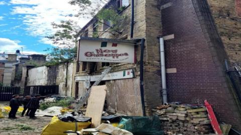 Three men stand outside a crumbling stone building with a sign on the wall that reads 'New Giuseppes'