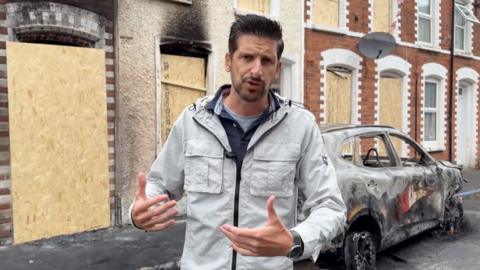 Correspondent Dan Johnson reporting from Belfast street where a burnt car is seen