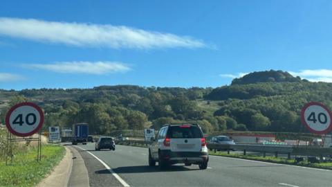 A stream of traffic passes through a section of dual carriageway with signs saying that the road is limited to 40 miles per hour. Further back there are more signs letting drivers know that there are speed cameras in place. There is a white car in the foreground, wit ha black car further down the road and two lorries in front of that.