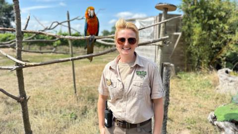 A woman wearing a safari outfit, sunglasses and with a walkie talkie on her belt, stands outside in a field while a parrot is perched on a branch behind her.