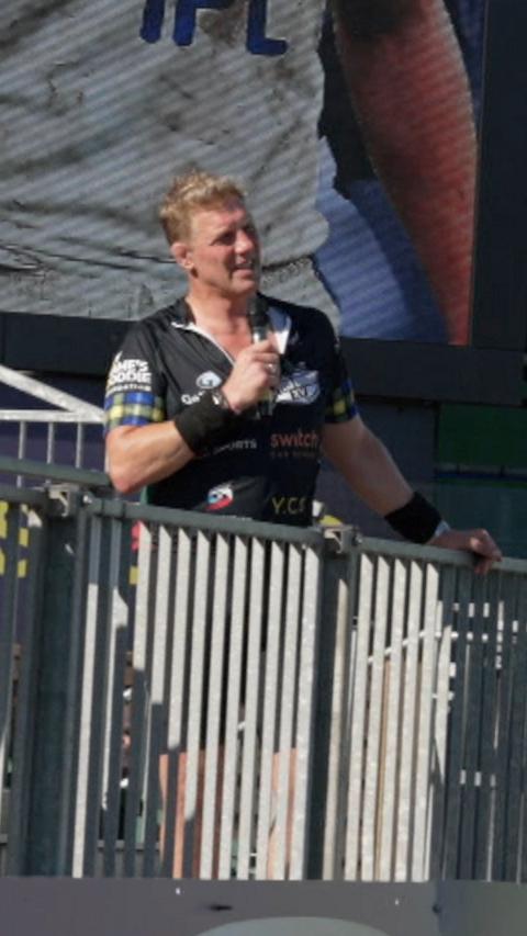 Former England captain Lewis Moody stands behind some railings on a stand at Bath Rugby's Recreation Ground. He's wearing a black cycling outfit with tartan sleeves and black sweatbadnds. He's holding a microphone. There is a big screen in the background showing a photograph of Lewis Moody playing for Bath.