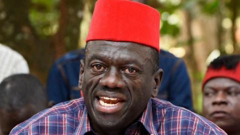 Kizza Besigye talks while wearing a red hat and multicoloured shirt.