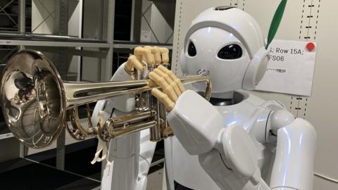 A white robot with large eyes and slightly human-like, with fingers holding a trumpet.