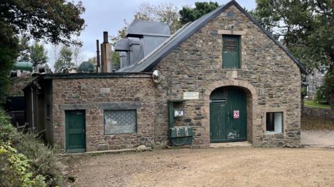 The Sark Electricity power station. A brown brick building, with a green door and a green petrol pump outside it. 

Two big metallic curved chimneys are on the roof, next to two metallic cylindrical chimneys.  