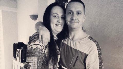 A black and white photo of a couple, David and his wife, smiling and hugging. In casual clothes at home