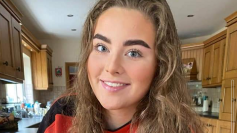 Jeni Larmour pictures in her kitchen, smiling wearing a red t-shirt.