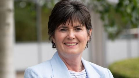 A head and shoulders shot of a woman with short dark hair and wearing a pale blue jacket, blue and white striped top and silver drop earrings. She is standing outside an office building with trees and grass visible in the blurred background. 