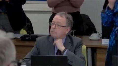 Richard Cook sat at a desk with a laptop during a council meeting