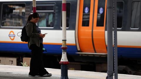 A woman standing on a London overground train platform wearing a grey jacket, black trousers and rucksack on her back looking at her mobile phone.