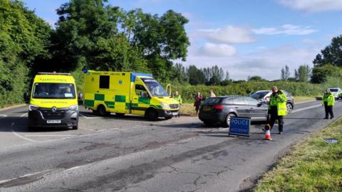 Two ambulances parked up after an accident at a crossroads. It is a rural road with hedgerows and grass banks either side. There is a "police accident" sign, with the text in white on a blue background, with police tape across the road