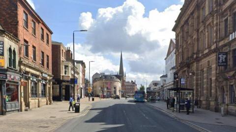 A high street in Wakefield, West Yorkshire, with stone buildings on either side and people walking on the pavements. There is a bus stop on one side with people waiting for a bus.