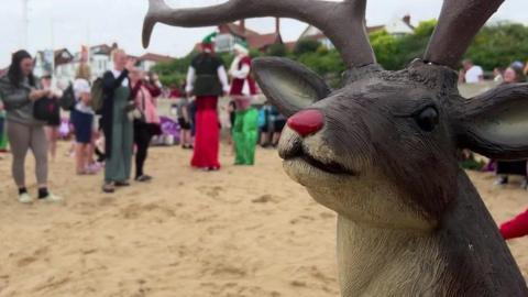 A life-sized model reindeer with a red nose stands on a beach with golden sand while stilt-walkers dressed as elves walk among spectators in the background, which is out of focus.
