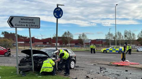 Police officers inspect a black car that has crashed into a road sign. Police officers are also visible in the background