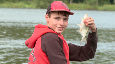 James Wellwood, wearing a red hoody and cap, looks towards the camera over his shoulder and holds up a smal fish