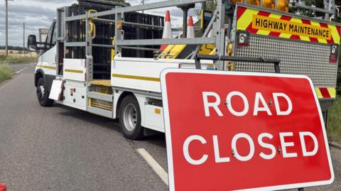 A road has been closed and has cones going across it. There is a sign in the middle of the road which is red and has ROAD CLOSED on it in white writing. A truck is parked across the road.