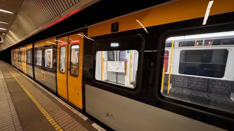A yellow, grey and black Metro train pulled into a station. The train is empty and its destination is Pelaw which is lit up on an LED display by the doors. The platform has a brown and yellow tiled floor.