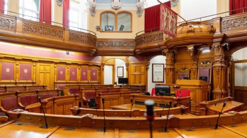 The interior of the States Assembly which is a round room lined with wooden benches with red leather backs that have desks in front of them. The second storey has windows with floor-length red curtains and the seat the Presiding Officer sits in features wooden carvings and has the Jersey flag hanging above it.