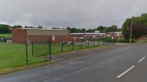 Red brick school buildings pictured from a distance. A road and green, metal fencing is in the foreground.