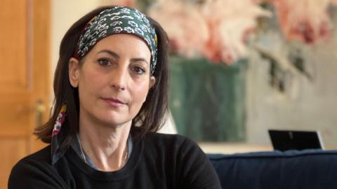 Donna looks at the camera as she sits on a blue sofa. She has a black jumper on and a head scarf with floral patterns on it. She has dark brown hair which comes down to her shoulders. Behind her on the wall is a painted flower pot with pink flowers inside.