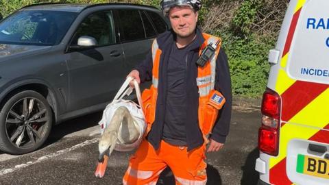 A man wearing orange high-vis uniform and a hard hat while holding an injured swan inside a specialist animal bag