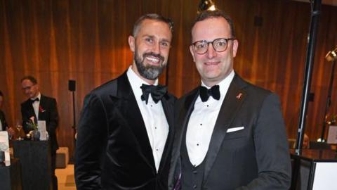 Jens Spahn (R) in glasses, a grey jacket and white shirt and bowtie beside his husband Daniel Funke who has as beard and a black velvet jacket with a white shirt and bowtie