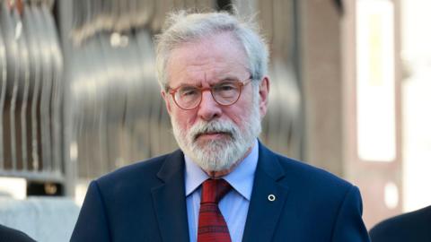 Gerry Adams walks outside the High Court in Dublin. He has short white hair and beard. He is wearing small, orange circular glasses and his expression is neutral. He is also wearing a navy suit jacket, a light blue collared shirt and a red patterned tie. On his suit jacket lapel he is wearing a silver fáinne, denoting his Irish speaking ability.