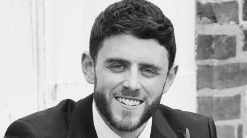 Andrew Harper is pictured in a black and white photograph on his wedding day, wearing a dark suit with a flower buttonhole. He has dark hair and a beard.
